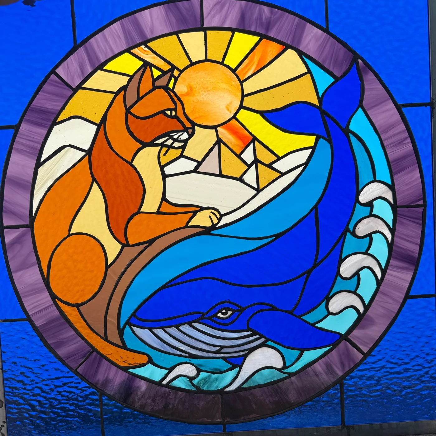 Cat & Whale: a round stained glass window of an orange cat on a branch above a blue whale in the waves, under a bright sun, framed in violet on cobalt glass